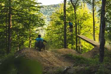 A mountain biker performing a jump off a ramp in a wooded area, while another rider in a wheelchair bike approaches a dirt mound. The scene is bathed in warm sunlight, surrounded by lush green trees and a hill in the background. The Driving Range mountain bike trail.