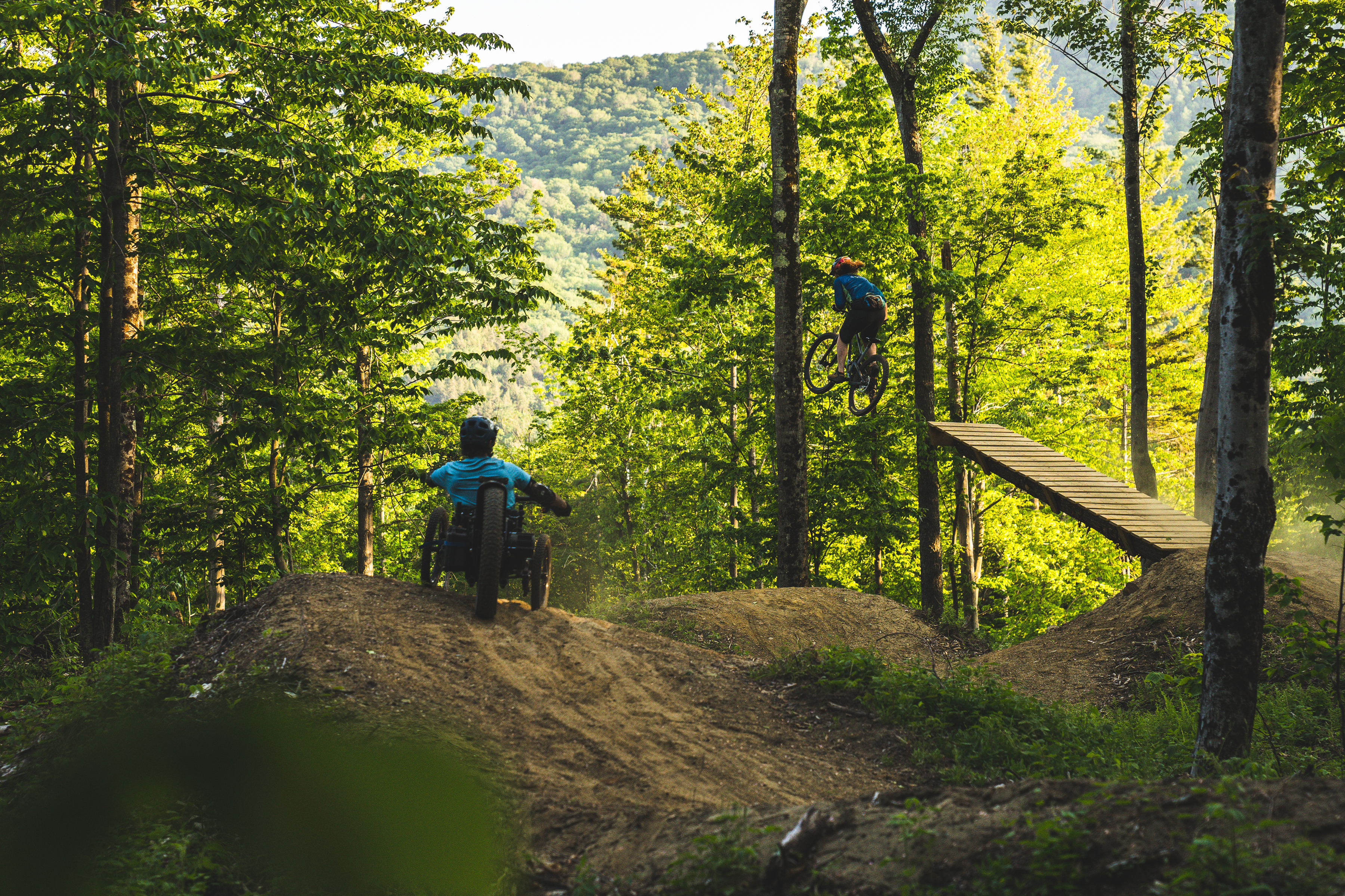 A mountain biker performing a jump off a ramp in a wooded area, while another rider in a wheelchair bike approaches a dirt mound. The scene is bathed in warm sunlight, surrounded by lush green trees and a hill in the background. The Driving Range mountain bike trail.