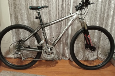Trek 4300: A silver and black mountain bike is positioned against a backdrop of light-colored curtains. The bike features a sleek frame, wide tires, and disc brakes, highlighting its design for off-road cycling. The seat is adjustable, and the handlebars have various controls attached, showcasing its functionality for cycling enthusiasts. The floor beneath the bike is wooden, adding a warm tone to the overall scene.
