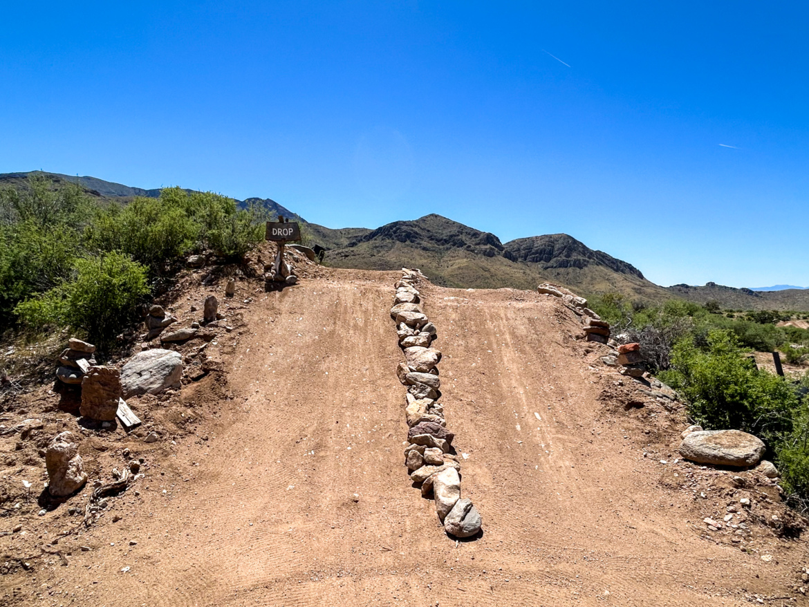 A dirt path leads to a steep drop-off, marked by a sign that reads "DROP." The surrounding landscape features rocky hills and sparse vegetation under a clear blue sky. Revenant Bike Park mountain bike trail.