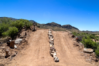 A dirt path leads to a steep drop-off, marked by a sign that reads "DROP." The surrounding landscape features rocky hills and sparse vegetation under a clear blue sky. Revenant Bike Park mountain bike trail.
