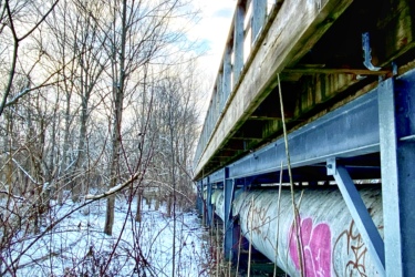 A view from underneath a wooden bridge, with metal support beams and a large pipe running underneath. The ground is covered in snow, and bare trees surround the scene, creating a wintry atmosphere. There is graffiti visible on the pipe, including a pink heart shape. The sky is partly cloudy, adding to the serene natural setting. MeadowLilly mountain bike trail.