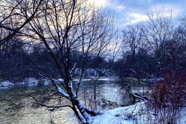 A tranquil winter scene featuring a snow-covered path alongside a river. Leafless trees line the banks, reflecting the soft light of a cloudy sky. The ground is blanketed in fresh snow, creating a serene and picturesque landscape. MeadowLilly mountain bike trail.