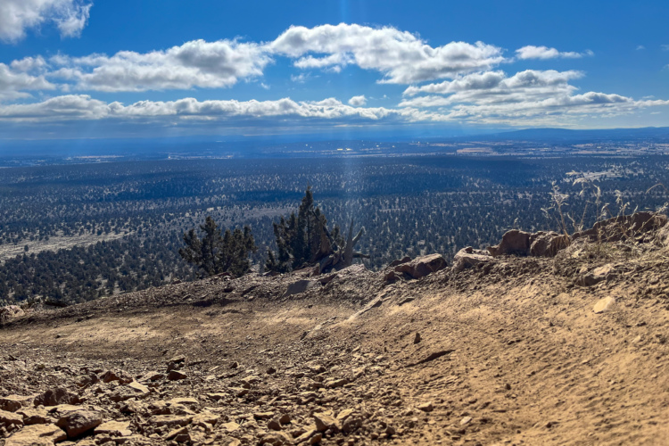 Wide view of a mountainous landscape under a blue sky with scattered clouds, featuring rugged terrain and distant tree-covered hills. The foreground shows a rocky path leading towards the horizon, showcasing the natural beauty of the area.