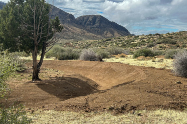 A natural landscape featuring a dirt bike ramp with an incline, surrounded by low shrubs and a single green tree on the left. The background showcases rolling hills and mountains under a cloudy sky. The ground is mostly bare soil with some patches of grass. Revenant Bike Park mountain bike trail.