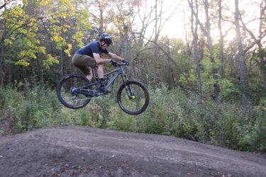 A person riding a mountain bike jumps off a dirt ramp in a wooded area, surrounded by trees with autumn foliage. The biker is wearing a helmet, a black shirt, and shorts, with colorful socks visible as they soar through the air. Fourmile Park mountain bike trail.