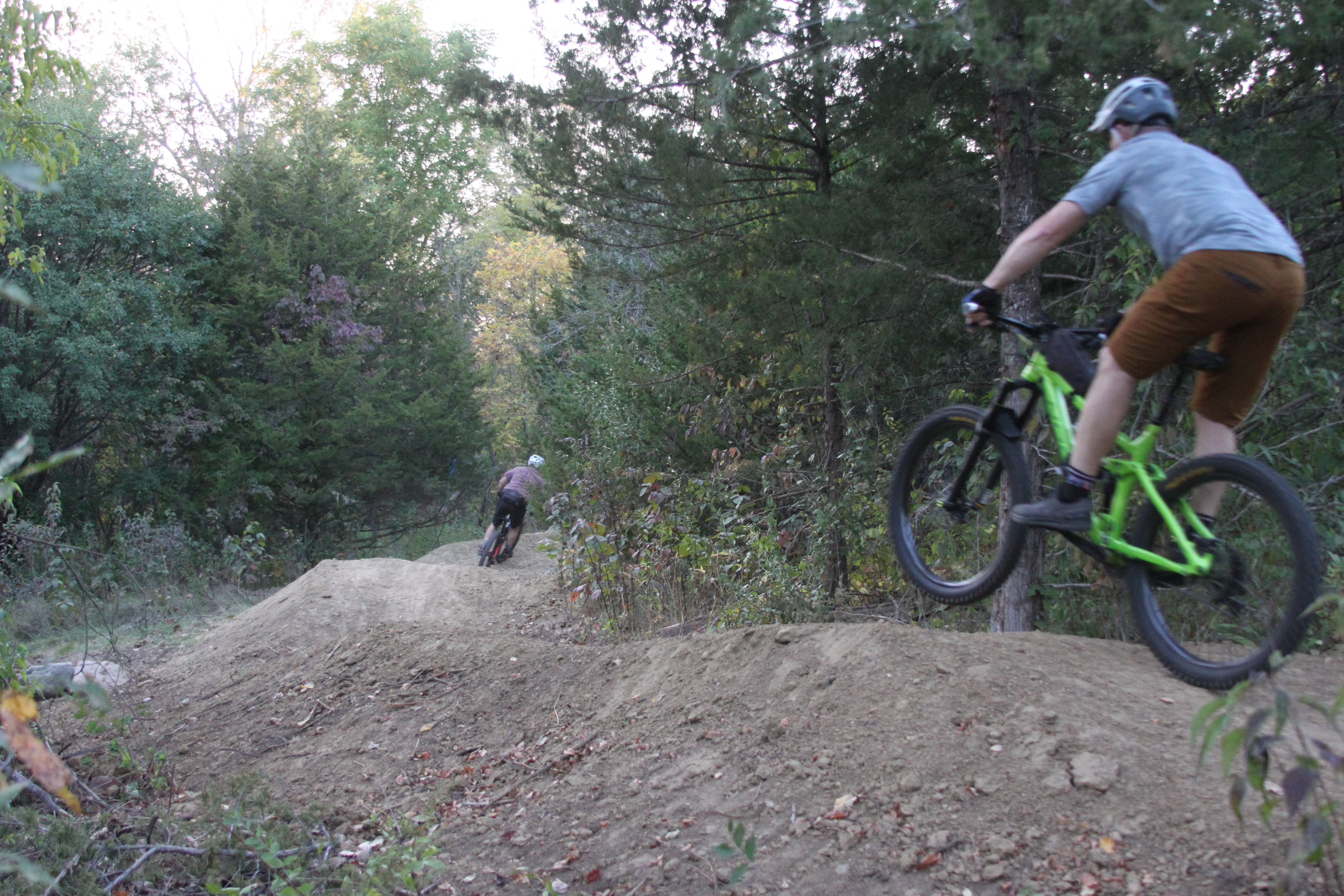 Two mountain bikers riding on a dirt trail in a forested area. One biker is airborne over a dirt jump, while the other is navigating a turn on the trail. The setting features trees and natural foliage, showcasing a vibrant outdoor environment. Fourmile Park mountain bike trail.