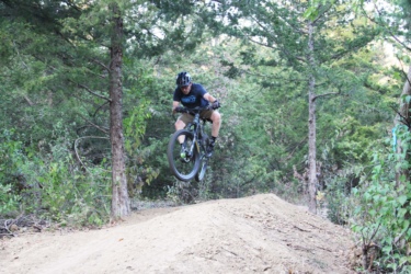 A cyclist performing a jump on a mountain bike over a dirt ramp, surrounded by trees and greenery in a natural outdoor setting. Fourmile Park mountain bike trail.