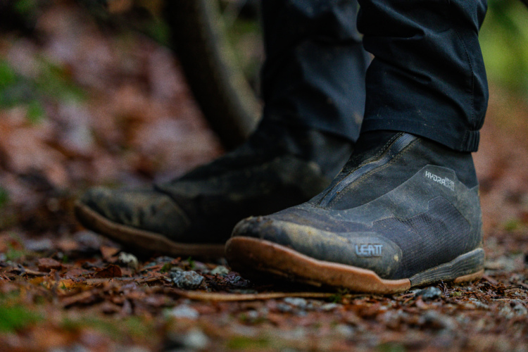 A close-up view of black waterproof biking shoes, partially covered in mud, positioned on a leaf-strewn trail. The shoes feature a sleek design with gray accents, showcasing the brand name "Leatt" on the side. The background is blurred, emphasizing the shoes against a natural, outdoor setting.