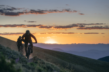 Two mountain bikers riding on a scenic trail at sunset. The sky is filled with colorful clouds and the sun is setting over distant mountains, providing a beautiful backdrop for their ride. The terrain is hilly with grassy patches, and the bikers are focused as they navigate the path. Toiyabe Crest Trail mountain bike trail.