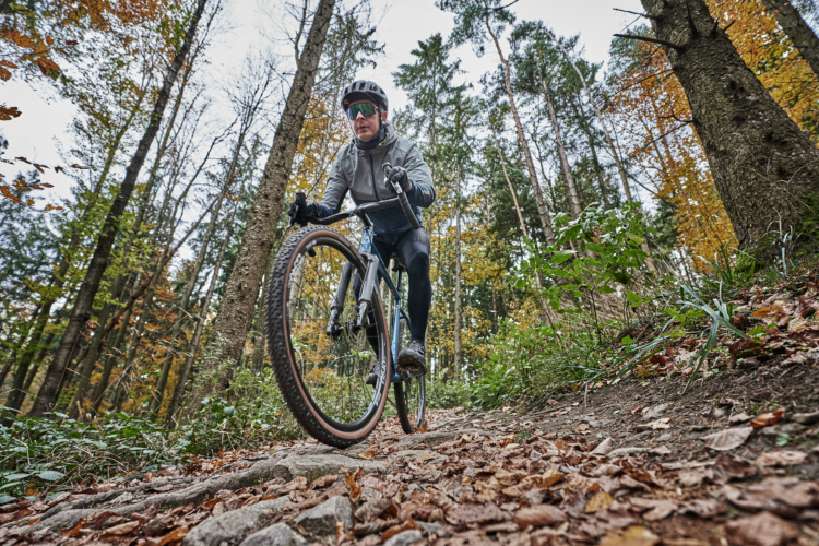 A person in cycling gear riding a Tout Terrain Ceres gravel bike on a rocky trail surrounded by autumn foliage and trees.