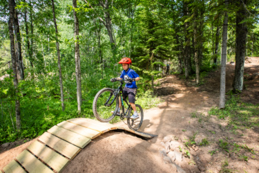 A young boy wearing a red helmet and a blue t-shirt rides a mountain bike over a wooden ramp in a dense forest area, surrounded by green trees and foliage. The path leads deeper into the woods, providing a sense of adventure. WinMan Trails mountain bike trail.