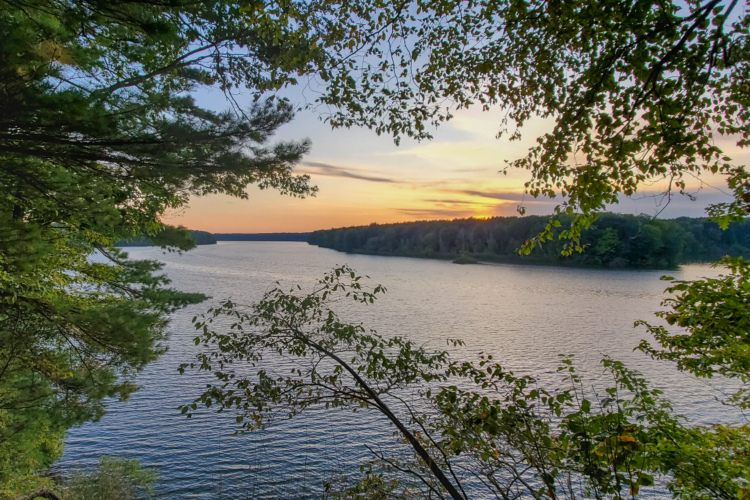 A serene view of a river framed by lush green trees, reflecting the colors of a sunset in the sky. The water is calm, with gentle ripples, and the landscape features wooded hills in the distance.