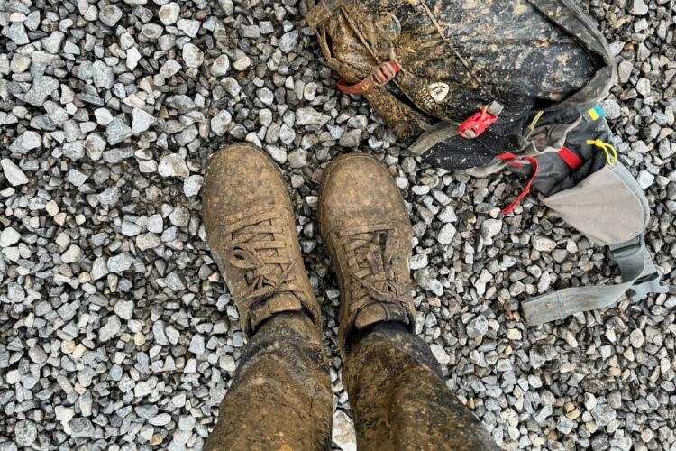 A pair of muddy shoes standing on a bed of gravel, with pants also covered in dirt. A backpack is visible nearby, showing signs of wear and dirt.