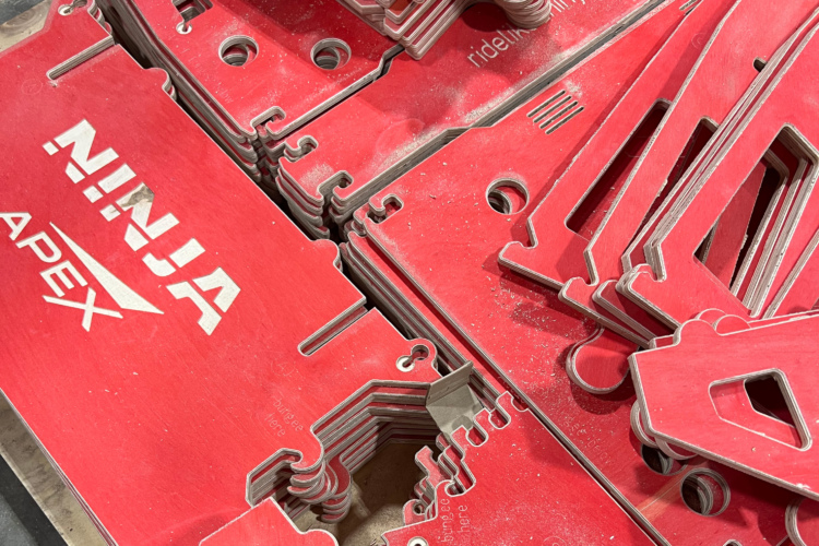 A collection of red acrylic sheets with various cut-out shapes, featuring the logos "NINJA" and "APEX." The surfaces of the sheets have a slight dusting, indicating they may have been recently manufactured or handled. The sheets are stacked together, showing their intricate designs and cut features.