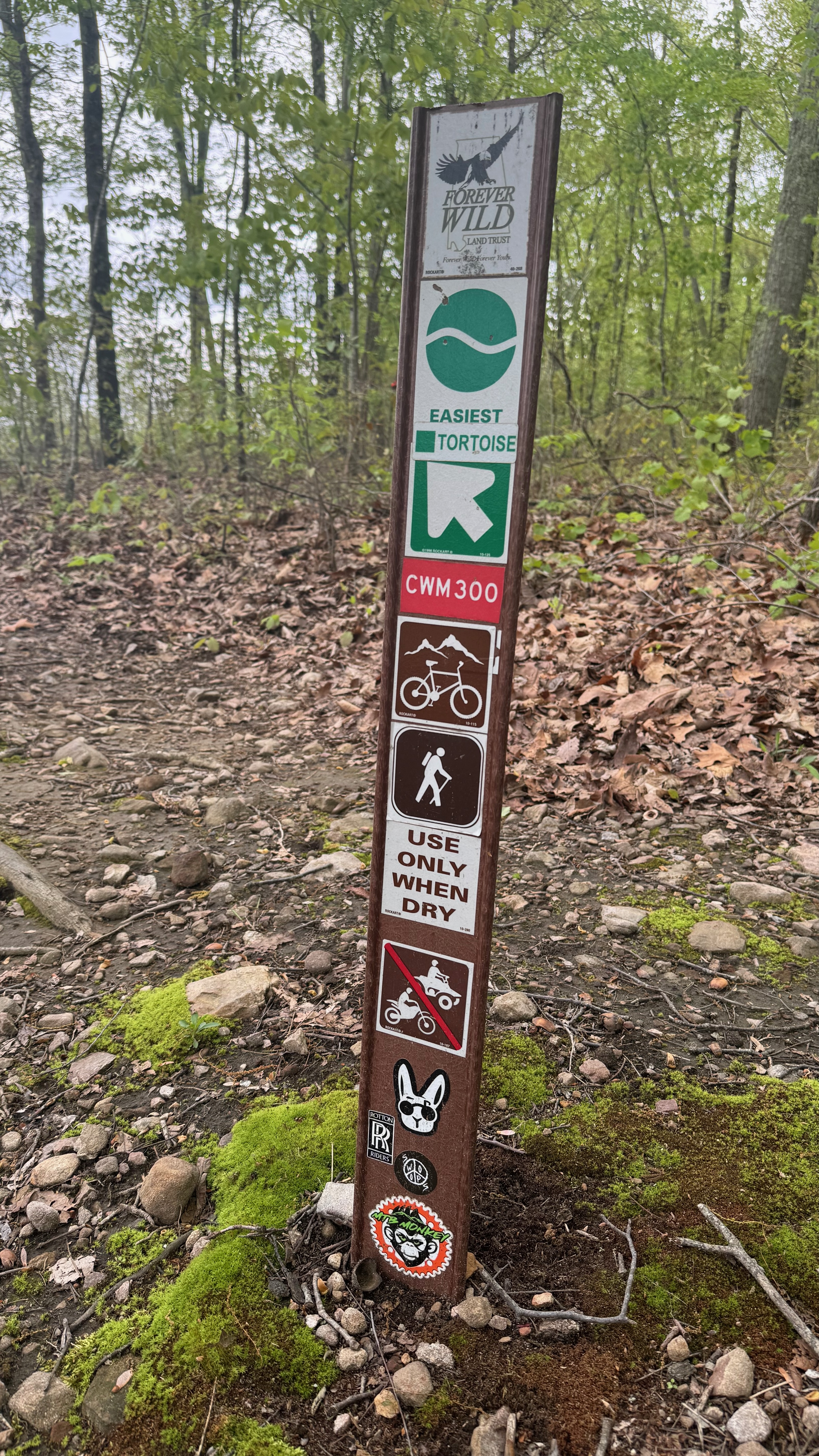 A trail marker sign indicating the "Easiest Tortoise" hiking trail in a wooded area. The sign features various symbols, including icons for hiking, biking, and a warning to "Use Only When Dry." There are also stickers at the bottom of the post. Surrounding the sign are green plants and fallen leaves on the ground. Tortoise mountain bike trail.