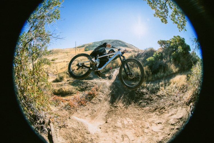 Cam Zink performing a jump over a dirt path on the Vacay, captured from a low angle with a fisheye lens. The background features rolling hills and clear blue skies, while the surrounding vegetation includes dry grass and sparse trees.