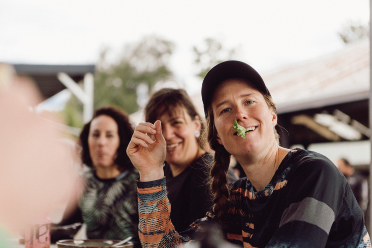 A group of people sitting together at a table, enjoying a meal outdoors. One woman in the foreground is smiling with a piece of greenery in her mouth, while others in the background appear engaged and smiling as well. The setting features a casual, relaxed atmosphere with greenery and rustic decor.