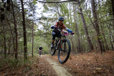 A mountain biker airborne over a dirt trail, surrounded by trees and foliage, with another cyclist following closely behind. The rider in the foreground wears a helmet and cycling gear, and has a race number displayed on their bike. The Ridgeland Trails mountain bike trail.