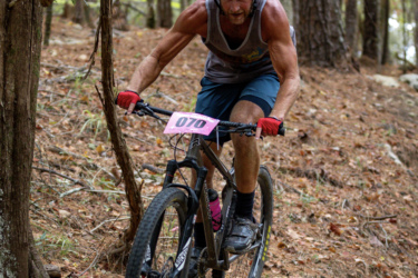 A muscular mountain biker in a red helmet navigates a rocky trail in a wooded area, focused on maintaining balance as he rides over a boulder. He is wearing a sleeveless shirt and shorts, with a race number displayed on the front of his bike. The forest is filled with trees and fallen leaves, creating a natural, rugged backdrop. The Ridgeland Trails mountain bike trail.
