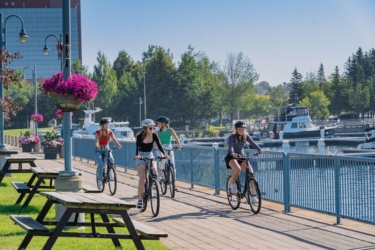 Five young adults are riding bicycles along a scenic pathway next to a marina, with boats docked in the background. The pathway features colorful hanging flower baskets and benches, while lush greenery and clear blue skies create a vibrant atmosphere. Hub Trail mountain bike trail.