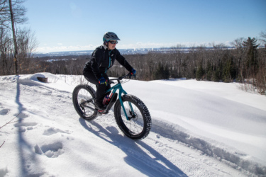 A person riding a fat bike on a snowy trail, surrounded by a winter landscape with trees and a clear blue sky. The cyclist is wearing a helmet, sunglasses, and winter clothing, navigating through the snow-covered terrain. Crimson Ridge mountain bike trail.