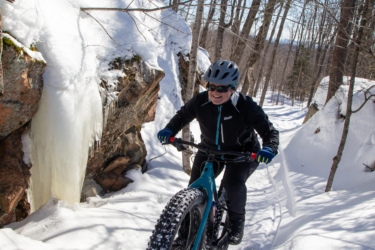 A person riding a fat bike on a snow-covered forest trail. The scene features trees in the background and a rocky formation with icicles hanging down. The cyclist is wearing a helmet and sunglasses, with a smile on their face, as they navigate through the snow. The bright blue sky indicates clear weather. Crimson Ridge mountain bike trail.
