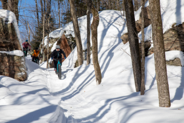 A group of four cyclists navigating a snow-covered trail surrounded by trees and rocks, with clear blue skies overhead. Crimson Ridge mountain bike trail.