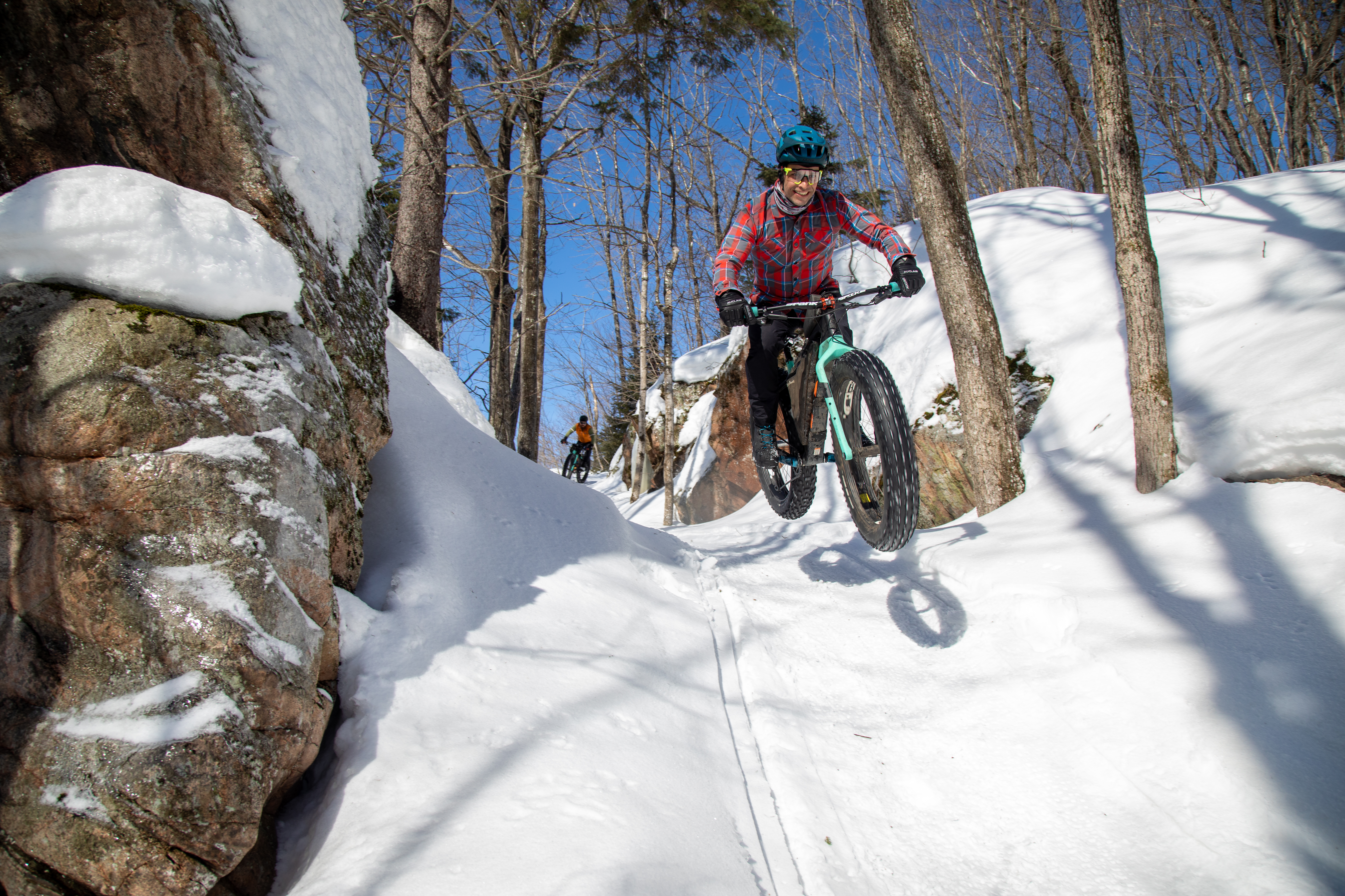 A cyclist jumps off a snowy trail surrounded by trees and rock formations, with another cyclist visible in the background. The scene is bright and clear, showcasing a winter biking adventure.