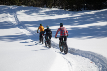 Three cyclists ride on a snow-covered path in a winter landscape, with tracks visible in the fresh snow. The cyclists wear winter attire and helmets, accompanied by tall trees in the background. The scene captures a bright, sunny day with clear blue skies. Crimson Ridge mountain bike trail.