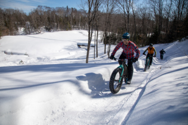 Three cyclists riding fat bikes on a snowy trail in a forested area. The sun is shining, and the ground is covered in fresh snow, creating a bright winter scene. The first cyclist, wearing a plaid shirt and helmet, is smiling as they navigate the trail, while two others follow behind. Trees are bare, indicating it is winter, and a snowy landscape stretches out in the background. Crimson Ridge mountain bike trail.