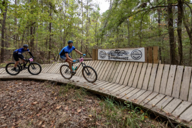 Two mountain bikers navigate a wooden track in a forested area, with a sign reading "Uncle Lumbuster" visible in the background. The trees are lush with green leaves, creating a vibrant, outdoor setting for the cyclists. The Ridgeland Trails mountain bike trail.