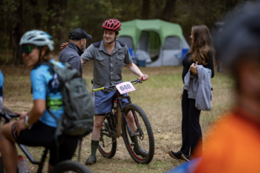 A group of mountain bikers gather at an outdoor event. A young man in a gray shirt and helmet smiles while talking to another man, standing by his bicycle with race number 040. Nearby, a woman in casual attire observes the interaction, while other cyclists are visible in the foreground. Tents are set up in the background among the trees. The Ridgeland Trails mountain bike trail.