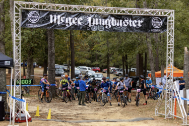 A group of mountain bikers gathered at the starting line of the McGee's Lungbuster event. The scene features a banner overhead, with a digital timer displaying the countdown. Participants are preparing for the race, while spectators and vehicles are visible in the background surrounded by trees. The Ridgeland Trails mountain bike trail.