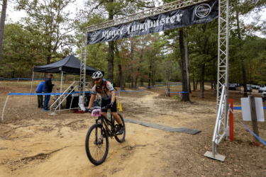 A cyclist wearing a helmet and race number 059 rides over a dirt path under a banner that reads "Jungle Buster." In the background, there is a black tent with people gathered, and trees line the trail, creating a natural setting for the event. The Ridgeland Trails mountain bike trail.