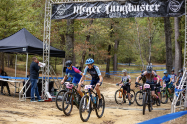 A group of mountain bikers compete in a race under a banner labeled "McGee Lungbuster." The scene shows several riders wearing colorful jerseys and helmets as they navigate a dirt path. In the background, a spectator area is visible, with a person speaking into a megaphone and others sitting nearby. The setting is surrounded by trees, indicating an outdoor trail event. The Ridgeland Trails mountain bike trail.