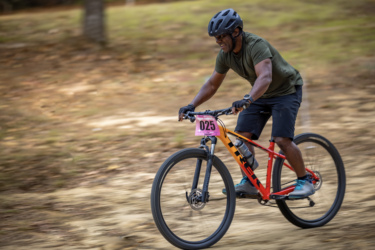 A person riding a mountain bike on a dirt path, wearing a black helmet and a green shirt, with a number "025" displayed on their bike. The rider appears focused and is smiling as they navigate through a natural, wooded landscape. The Ridgeland Trails mountain bike trail.