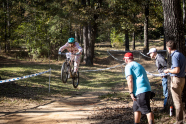 A cyclist in a colorful outfit jumps over a small mound on a dirt trail during a mountain biking event, while three spectators watch attentively from the sidelines in a wooded area. The Ridgeland Trails mountain bike trail.
