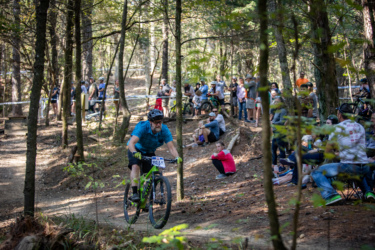 A mountain biker in a blue plaid shirt rides along a dirt trail through a wooded area, while a crowd of spectators watches from the side. The scene captures the excitement of a biking event, with trees surrounding the trail and sunlight filtering through the leaves. The Ridgeland Trails mountain bike trail.