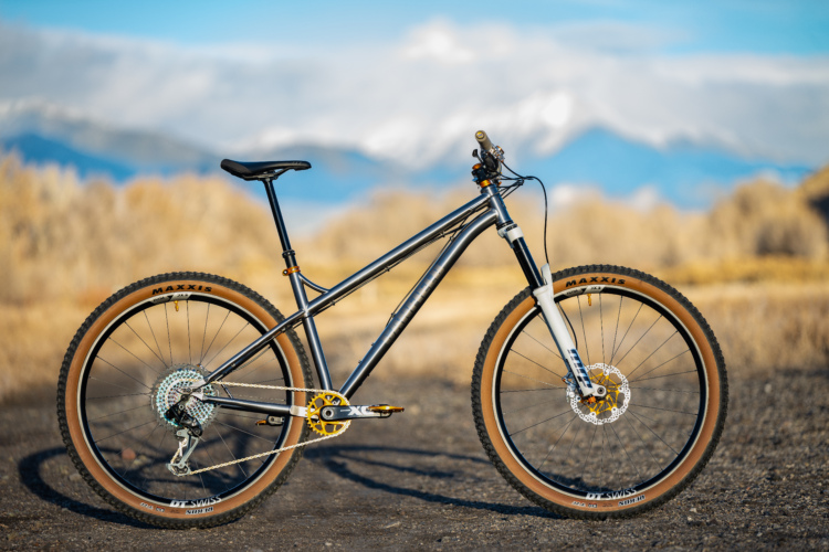 A mountain bike with a sleek silver frame and brown tires is positioned on a gravel surface. The bike features a single front suspension fork, a modern gear system, and a well-defined seat. The backdrop includes softly blurred mountains and grassy terrain, suggesting an outdoor setting ideal for cycling.
