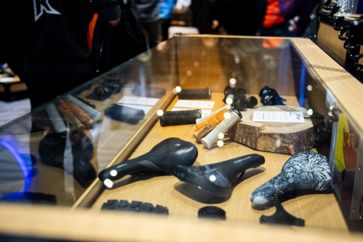 A display case featuring various bicycle components, including seats, grips, and accessories. The items are arranged on a wooden surface, with several tags providing information. Reflections and surrounding figures are visible in the glass.