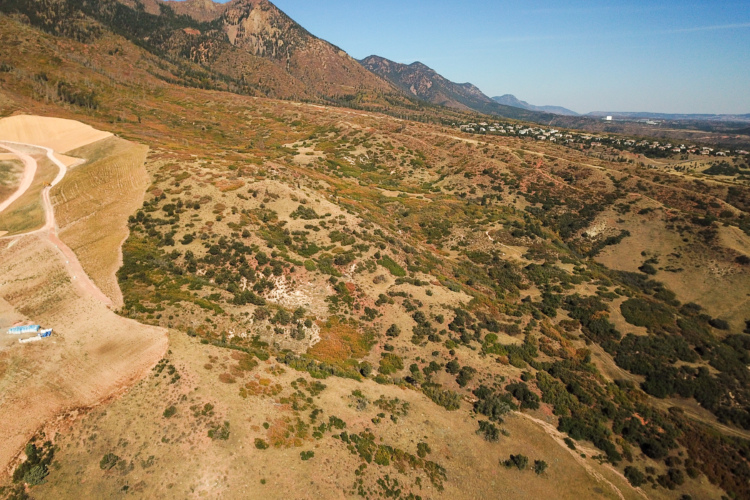 Aerial view of a mountainous landscape featuring rolling hills and sparse vegetation, with a dirt path winding through the terrain. The background showcases a range of mountains under a clear blue sky, while a small construction area with blue equipment is visible in the foreground. The scene conveys a sense of natural beauty and open space.