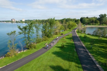 A scenic view of a bike path alongside a calm lake, with a lush green landscape and trees. Several people are riding bicycles along the path, which winds through a park-like setting, offering glimpses of the water and distant buildings against a clear blue sky. Hub Trail mountain bike trail.