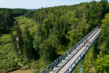 Aerial view of a long, elevated wooden bridge winding through a lush green forest. Several people are biking and walking on the bridge, surrounded by dense trees and a meandering stream below. The scene captures a serene outdoor atmosphere on a clear day. Hub Trail mountain bike trail.