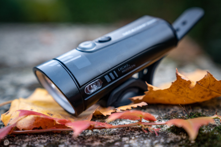 A close-up image of a black bicycle light resting on a textured surface, surrounded by autumn leaves. The light features a clear lens, buttons on the side, and markings indicating its brand. The warm tones of the fallen leaves contrast with the sleek design of the light.