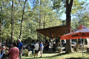 A lively outdoor gathering in a wooded area with a stage setup. People of various ages are socializing in the foreground, with some seated and others standing. A brightly colored tent featuring the "Kona" logo is visible, along with musical equipment on the stage. Tall trees and clear blue skies provide a picturesque backdrop, suggesting a festive atmosphere. The Ridgeland Trails mountain bike trail.