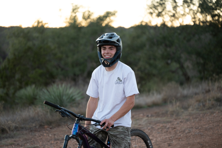 A young man wearing a helmet and a white t-shirt stands next to a mountain bike on a dirt trail, smiling at the camera. The background features lush greenery and a sunset sky, creating an outdoor atmosphere.