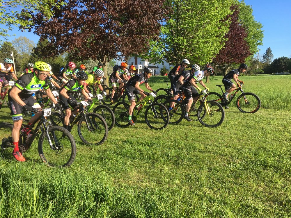 A group of mountain bikers lined up at the start of a race, ready to take off on a grassy field surrounded by trees. The cyclists, wearing colorful jerseys and helmets, are on their bikes with determination, prepared for the upcoming competition.