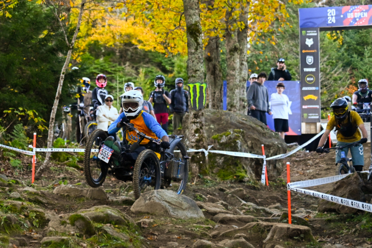 A mountain biker navigating a rocky trail during a race, with spectators in the background. The scene is set in a forested area with autumn foliage. The cyclist, in a blue and orange jersey, rides a specialized bike designed for off-road terrain. Nearby, a group of onlookers watches the event, with a race banner visible in the background.