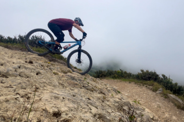 A mountain biker navigating a rocky terrain on a foggy day, leaning forward for balance as he rides down a steep section of the trail. Lush greenery is visible in the background, partially obscured by mist.